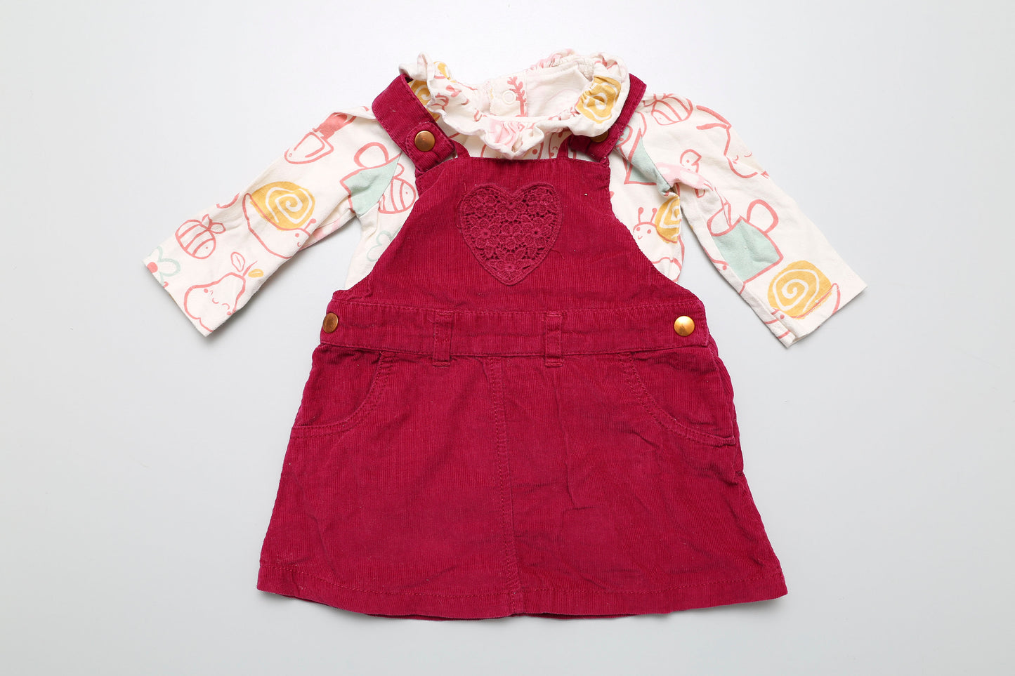 Buntes Baumwoll-Set von George. Das langärmelige Poloshirt hat ein fröhliches Tier- und Fruchtmuster, während das dunkelrosarote-rotstichige Latzkleid mit kleinen Herzen verziert ist. Größe 62-68.