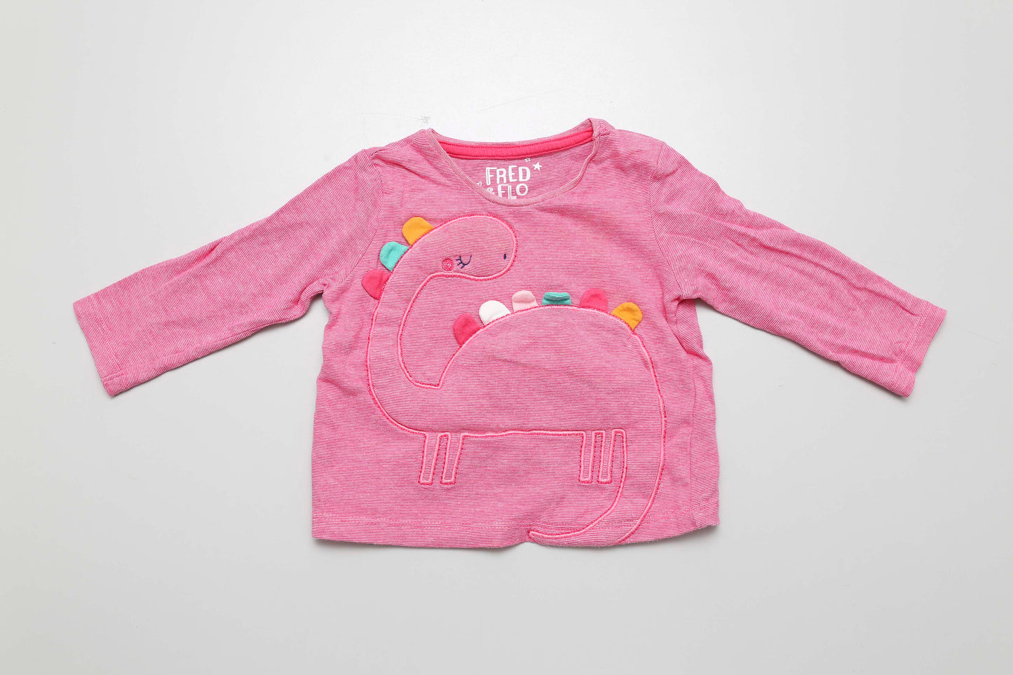Leuchtend rosa Langarmshirt mit Dinosauriermotiv. 100 % Baumwolle, Größe 68. Die Ärmel sind leicht verdreht.