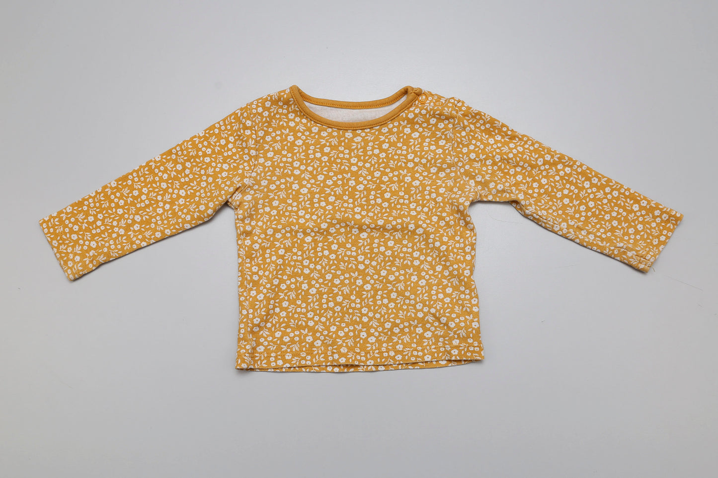 Fröhliches gelbes Langarmshirt mit zartem Blumenprint für kleine Mädchen. Aus 100 % Baumwolle, mit praktischen Druckknöpfen an der Schulter – angenehm weich, bequem und ideal für Größe 68/74.